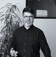 Black and white image of Damian standing looking directly to camera, wearing a black shirt with black framed glasses